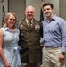 Three individuals pose together for a photo at what appears to be a military ceremony or formal event. The central figure is a U.S. Army officer in dress uniform with numerous medals and ribbons, flanked by a woman in a blue striped dress and a man in a casual blue button-down shirt. American flags are visible in the background.
