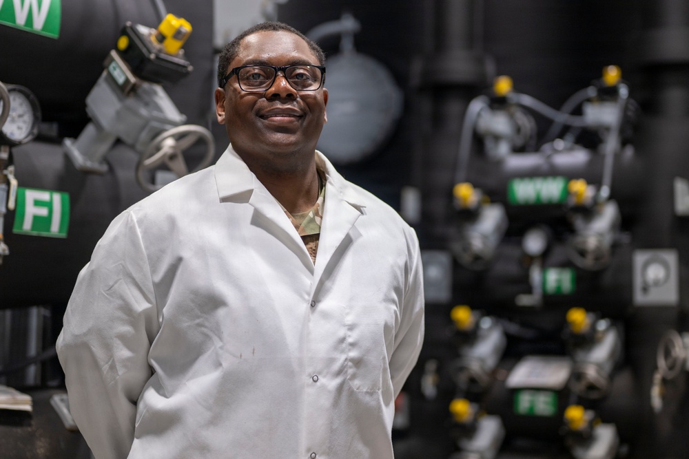 A man wearing a white lab coat and black-framed glasses stands confidently in what appears to be an industrial or mechanical facility. He is smiling and has his hands behind his back, posing for what appears to be a professional portrait. Beneath his lab coat, a camouflage uniform is partially visible at the collar.