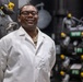 A man wearing a white lab coat and black-framed glasses stands confidently in what appears to be an industrial or mechanical facility. He is smiling and has his hands behind his back, posing for what appears to be a professional portrait. Beneath his lab coat, a camouflage uniform is partially visible at the collar.