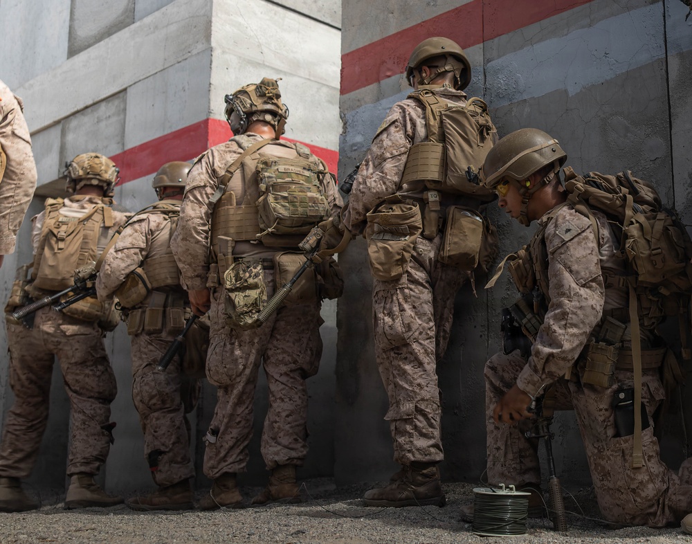 A group of military personnel in desert camouflage uniforms are positioned against a concrete wall, appearing to be in a tactical formation. One soldier is kneeling on the ground handling what appears to be a spool of wire or cable. The setting appears to be a training exercise or operational scenario based on the painted wall markings and equipment visible.