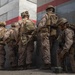 A group of military personnel in desert camouflage uniforms are positioned against a concrete wall, appearing to be in a tactical formation. One soldier is kneeling on the ground handling what appears to be a spool of wire or cable. The setting appears to be a training exercise or operational scenario based on the painted wall markings and equipment visible.