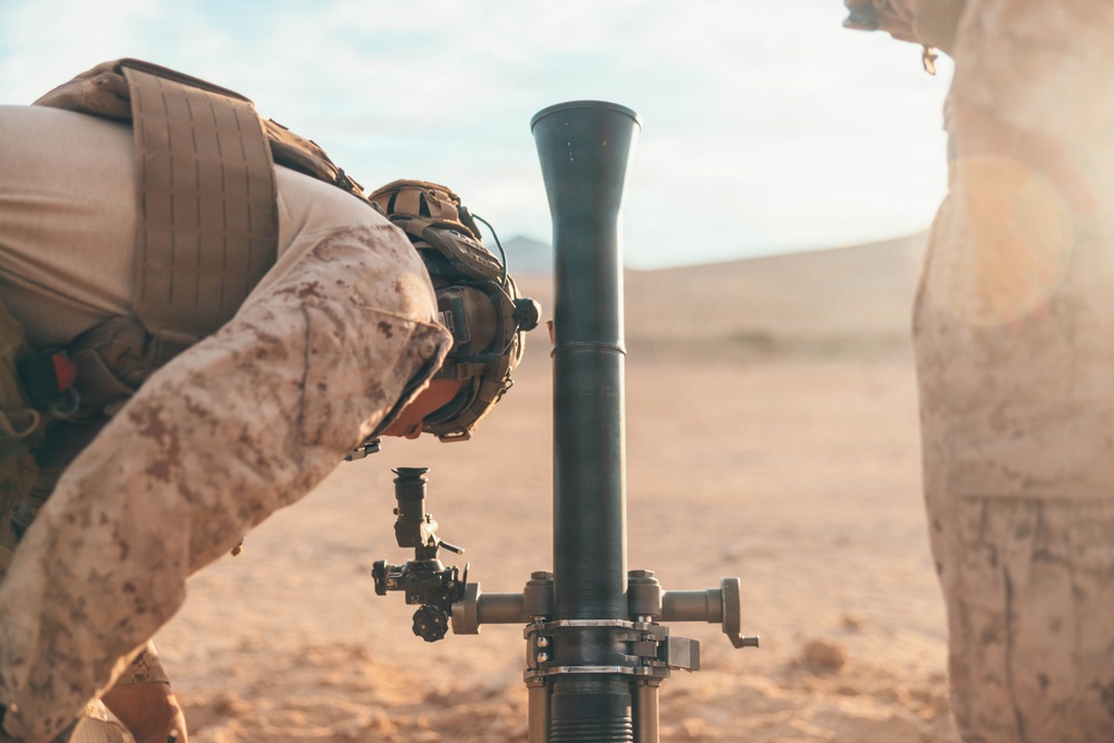 A soldier in desert camouflage uniform is leaning over to inspect or aim a mortar tube mounted on a bipod in an arid, desert environment. The mortar system is positioned vertically with its barrel pointing upward and a sight/aiming device attached to the side. A second individual in tan/desert clothing is partially visible on the right edge of the frame.