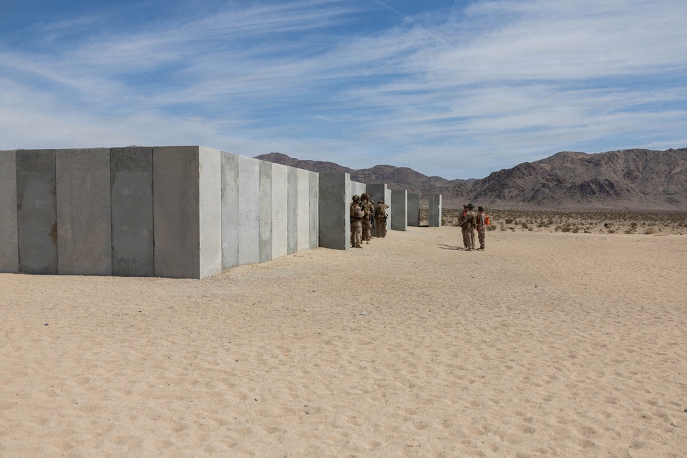 Military personnel in desert camouflage uniforms are conducting a training exercise at what appears to be a desert training facility, likely in the American Southwest. Large concrete wall panels are arranged in a corridor or maze-like configuration, suggesting urban warfare or close-quarters combat training. The arid landscape, sandy terrain, and mountain backdrop are consistent with locations such as the Marine Corps Air Ground Combat Center at Twentynine Palms, California.