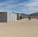 Military personnel in desert camouflage uniforms are conducting a training exercise at what appears to be a desert training facility, likely in the American Southwest. Large concrete wall panels are arranged in a corridor or maze-like configuration, suggesting urban warfare or close-quarters combat training. The arid landscape, sandy terrain, and mountain backdrop are consistent with locations such as the Marine Corps Air Ground Combat Center at Twentynine Palms, California.