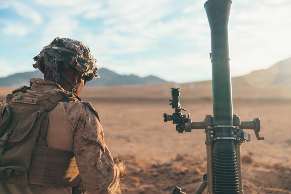 A military service member in desert camouflage uniform is positioned beside a mortar system in an arid, desert training environment. The soldier, identified by a name tape reading 'TOBARCHACON,' is wearing a tactical vest and a camouflage-covered helmet. The setting appears to be a desert training range with mountains visible in the background.