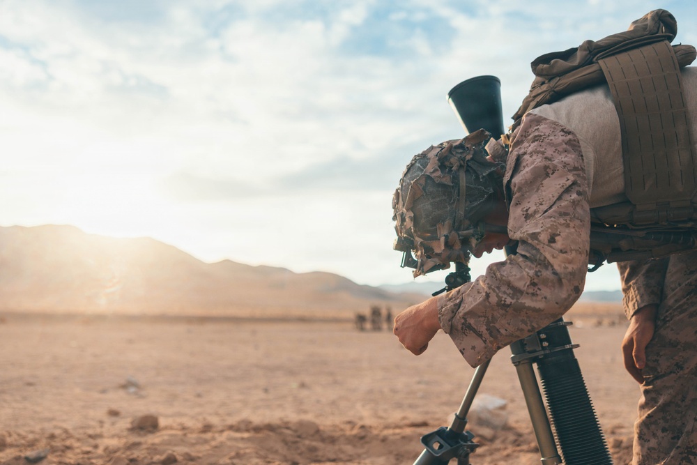 A soldier in desert camouflage uniform is operating what appears to be a mortar system mounted on a bipod in an arid, desert environment. The soldier is wearing full combat gear including a tactical vest, camouflage helmet with burlap cover, and is leaning over the weapon system. The background shows a flat desert landscape with distant mountains and partial cloud cover with visible sunlight.