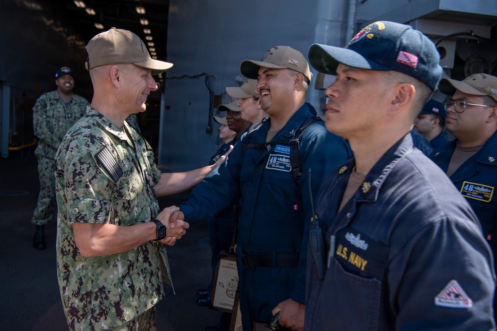 A senior U.S. Navy officer in digital camouflage (NWU Type III) is shaking hands with enlisted sailors dressed in Navy Working Uniform (NWU Type I, blue). The setting appears to be aboard a U.S. Navy vessel, likely on a hangar or flight deck. The interaction suggests an official visit, inspection, or recognition ceremony.