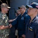 A senior U.S. Navy officer in digital camouflage (NWU Type III) is shaking hands with enlisted sailors dressed in Navy Working Uniform (NWU Type I, blue). The setting appears to be aboard a U.S. Navy vessel, likely on a hangar or flight deck. The interaction suggests an official visit, inspection, or recognition ceremony.