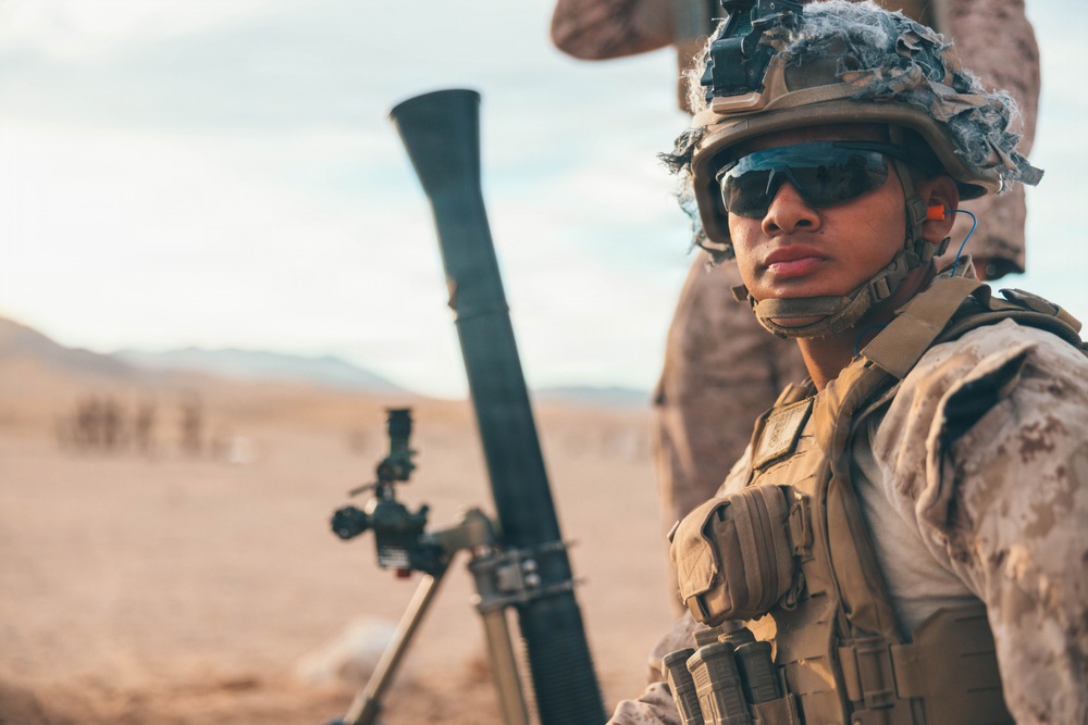 A soldier in full combat gear is positioned near a mortar system in an arid desert environment. The individual is wearing MARPAT (Marine pattern) camouflage uniform, tactical vest, combat helmet with netting, and dark ballistic sunglasses. The setting appears to be a training exercise or operational deployment in a desert region.