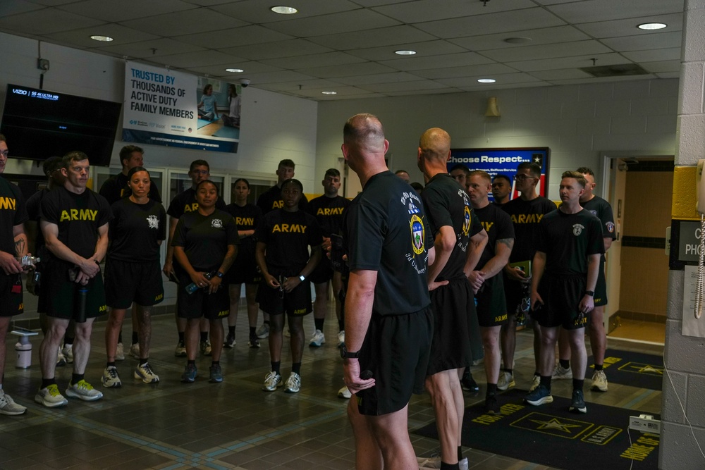 A group of U.S. Army personnel in black PT (Physical Training) uniforms are assembled indoors, likely in a fitness facility or gymnasium. Two individuals with their backs to the camera appear to be addressing or briefing the group. The setting suggests a pre- or post-workout formation or briefing session.