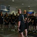 A group of U.S. Army personnel in black PT (Physical Training) uniforms are assembled indoors, likely in a fitness facility or gymnasium. Two individuals with their backs to the camera appear to be addressing or briefing the group. The setting suggests a pre- or post-workout formation or briefing session.