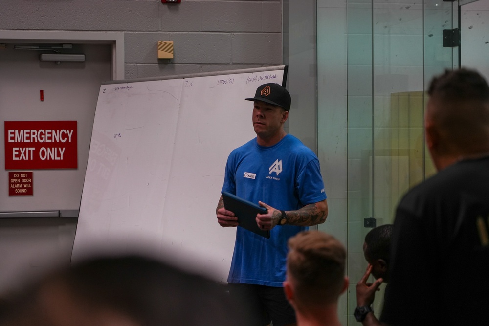 A man wearing a blue 'Apex Path' branded shirt and black cap is presenting or instructing a group of people in what appears to be an indoor facility. He is holding a tablet device and standing in front of a whiteboard with writing on it. An 'Emergency Exit Only' sign is visible on the wall to his left.