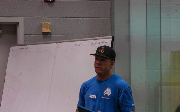A man wearing a blue 'Apex Path' branded shirt and black cap is presenting or instructing a group of people in what appears to be an indoor facility. He is holding a tablet device and standing in front of a whiteboard with writing on it. An 'Emergency Exit Only' sign is visible on the wall to his left.