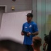 A man wearing a blue 'Apex Path' branded shirt and black cap is presenting or instructing a group of people in what appears to be an indoor facility. He is holding a tablet device and standing in front of a whiteboard with writing on it. An 'Emergency Exit Only' sign is visible on the wall to his left.