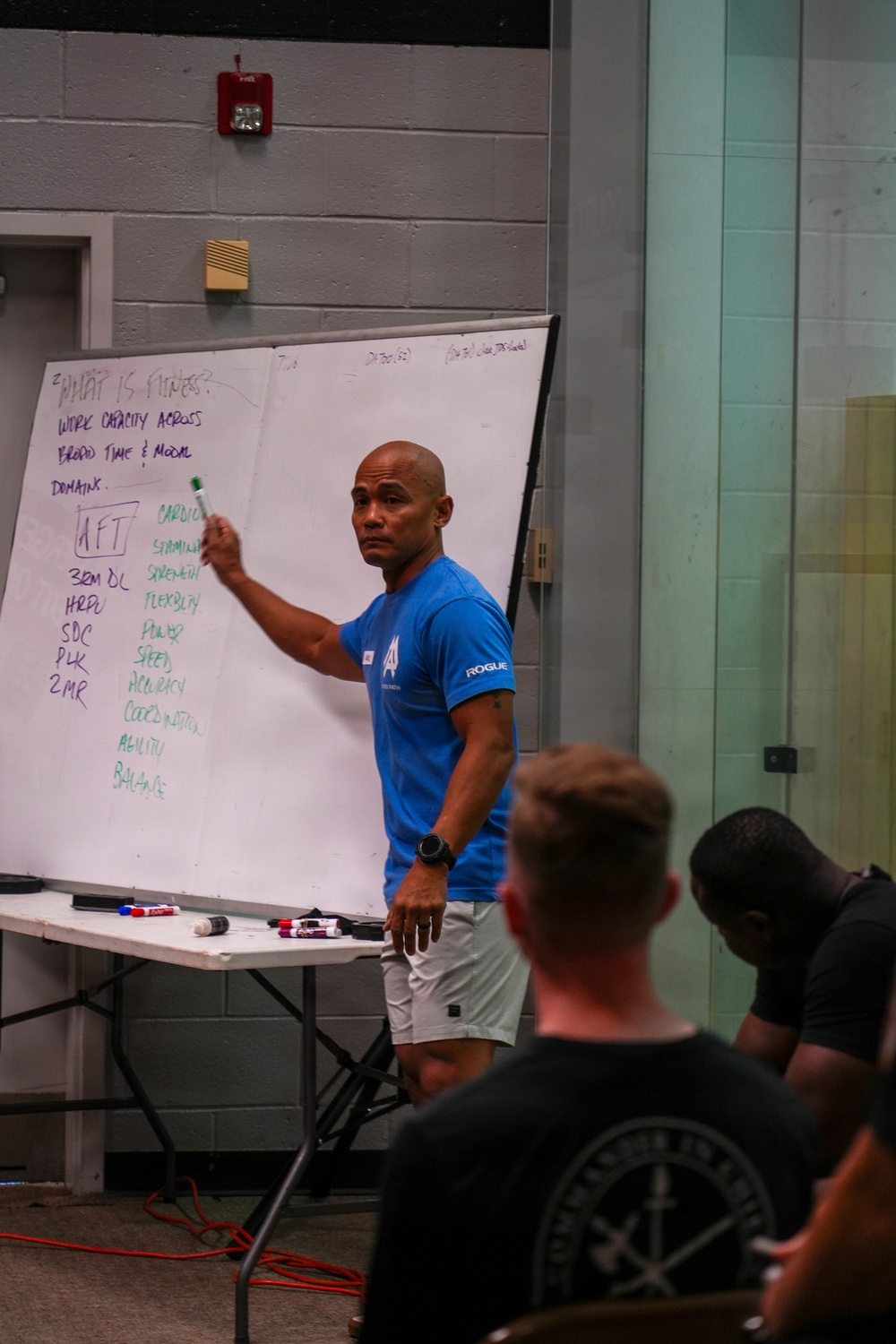 A fitness instructor or coach is presenting at a whiteboard during what appears to be a fitness education seminar or training session. The whiteboard displays the heading 'What is Fitness?' with content about work capacity, broad time and modal domains, and a list of physical fitness components. An audience of at least two people is visible in the foreground, seated and facing the presenter.