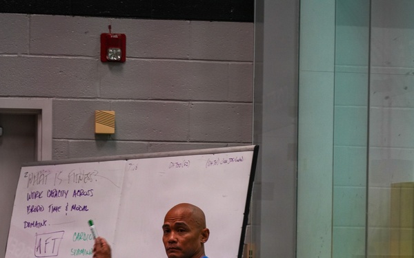 A fitness instructor or coach is presenting at a whiteboard during what appears to be a fitness education seminar or training session. The whiteboard displays the heading 'What is Fitness?' with content about work capacity, broad time and modal domains, and a list of physical fitness components. An audience of at least two people is visible in the foreground, seated and facing the presenter.