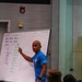 A fitness instructor or coach is presenting at a whiteboard during what appears to be a fitness education seminar or training session. The whiteboard displays the heading 'What is Fitness?' with content about work capacity, broad time and modal domains, and a list of physical fitness components. An audience of at least two people is visible in the foreground, seated and facing the presenter.