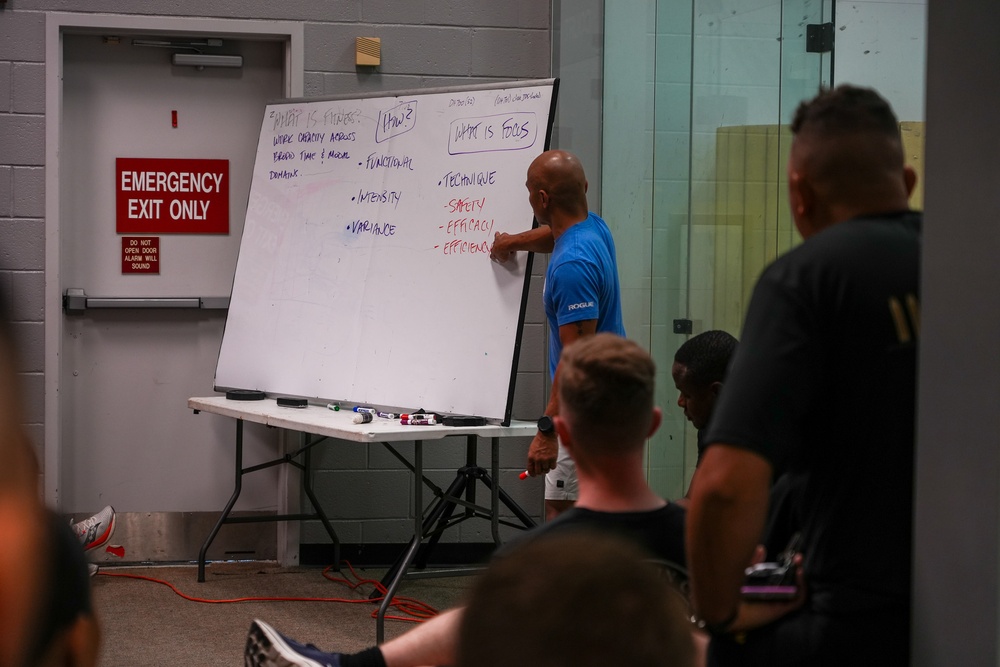 A fitness instructor in a blue shirt is presenting at a large whiteboard to a group of attendees in what appears to be a gym or training facility. The whiteboard contains notes about fitness concepts including definitions, focus areas, and key principles. The setting suggests a CrossFit or physical training seminar or certification course.