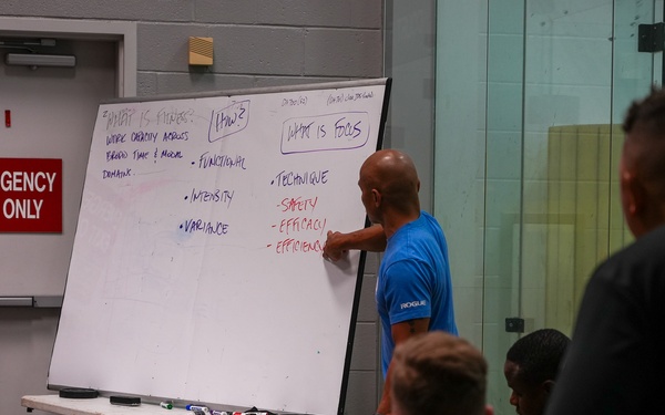 A fitness instructor in a blue shirt is presenting at a large whiteboard to a group of attendees in what appears to be a gym or training facility. The whiteboard contains notes about fitness concepts including definitions, focus areas, and key principles. The setting suggests a CrossFit or physical training seminar or certification course.