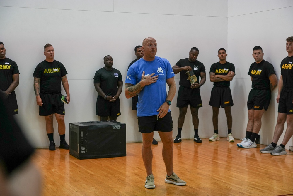 A civilian instructor or trainer in a blue shirt is addressing a group of U.S. Army soldiers in a gymnasium or indoor training facility. The soldiers are wearing standard black Army PT (Physical Training) uniforms and are standing along the walls listening attentively. A plyo box is visible on the floor, suggesting a fitness or physical training session is taking place.