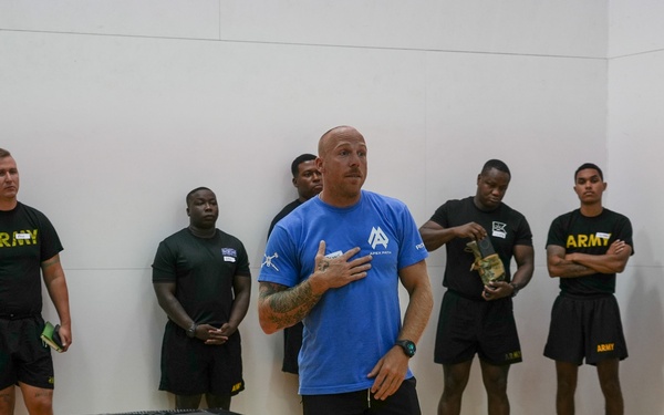 A civilian instructor or trainer in a blue shirt is addressing a group of U.S. Army soldiers in a gymnasium or indoor training facility. The soldiers are wearing standard black Army PT (Physical Training) uniforms and are standing along the walls listening attentively. A plyo box is visible on the floor, suggesting a fitness or physical training session is taking place.