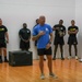 A civilian instructor or trainer in a blue shirt is addressing a group of U.S. Army soldiers in a gymnasium or indoor training facility. The soldiers are wearing standard black Army PT (Physical Training) uniforms and are standing along the walls listening attentively. A plyo box is visible on the floor, suggesting a fitness or physical training session is taking place.