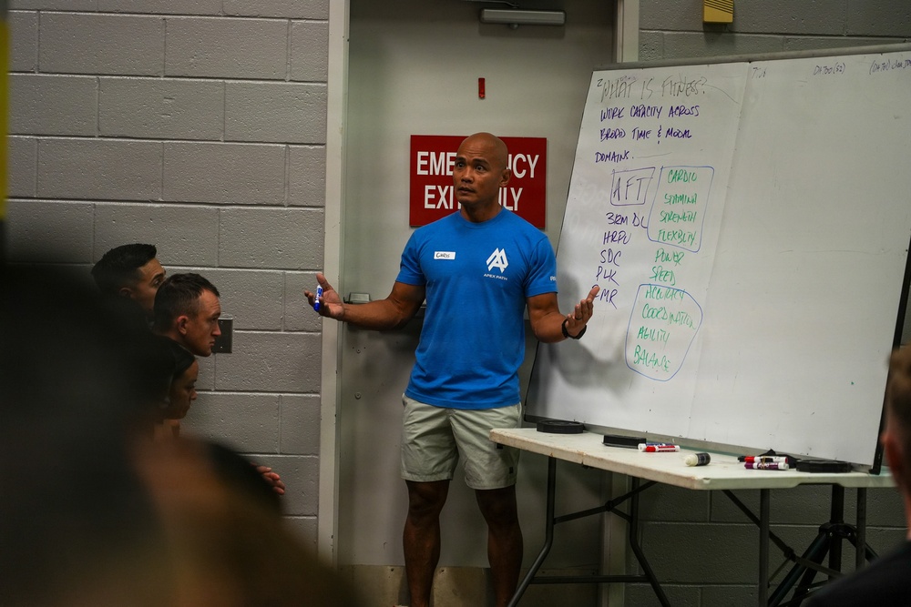 A fitness instructor wearing a blue 'Apex Parisi' t-shirt is presenting to a group of attendees in what appears to be a gym or training facility. He is standing in front of a whiteboard that displays notes about fitness definitions and components. The setting suggests a formal fitness education or coaching seminar.