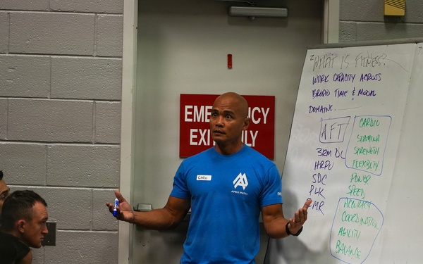 A fitness instructor wearing a blue 'Apex Parisi' t-shirt is presenting to a group of attendees in what appears to be a gym or training facility. He is standing in front of a whiteboard that displays notes about fitness definitions and components. The setting suggests a formal fitness education or coaching seminar.