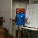 A fitness instructor wearing a blue 'Apex Parisi' t-shirt is presenting to a group of attendees in what appears to be a gym or training facility. He is standing in front of a whiteboard that displays notes about fitness definitions and components. The setting suggests a formal fitness education or coaching seminar.