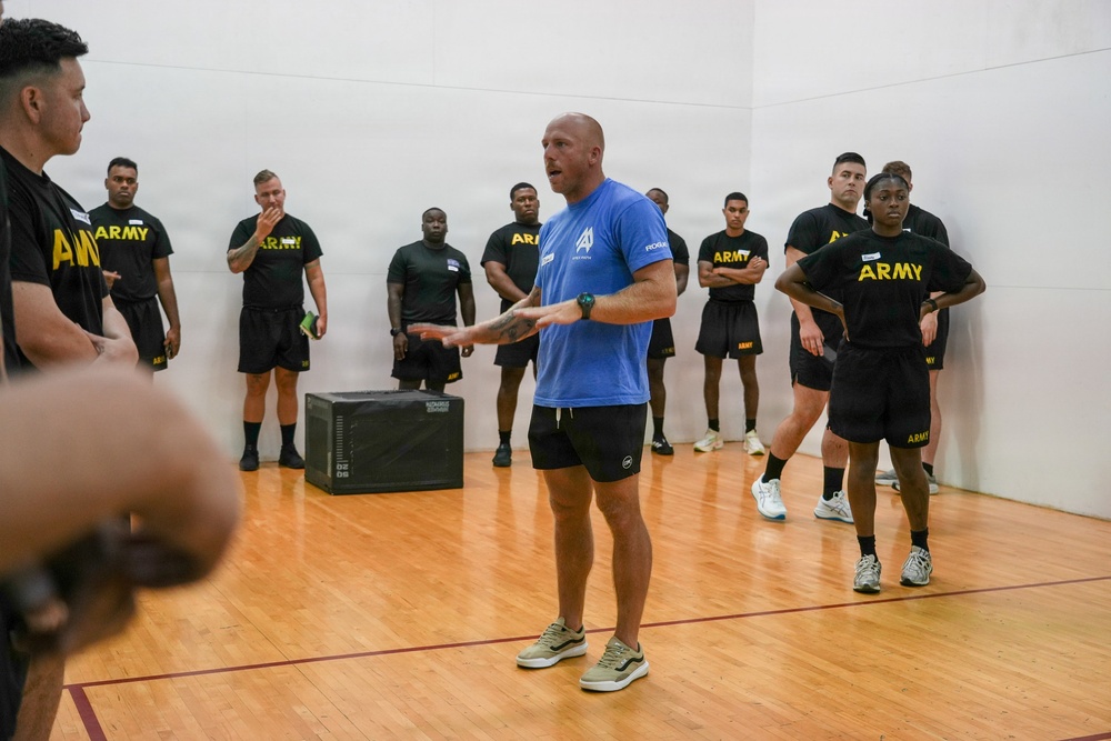 A civilian instructor or trainer in a blue shirt is addressing a group of U.S. Army soldiers wearing black 'ARMY' branded physical training uniforms inside what appears to be a gymnasium or racquetball court. The instructor appears to be demonstrating or explaining something with hand gestures while the soldiers stand attentively around him. A black plyo box is visible on the hardwood floor, suggesting a physical fitness or training session.