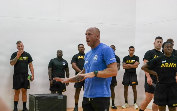 A civilian instructor or trainer in a blue shirt is addressing a group of U.S. Army soldiers wearing black 'ARMY' branded physical training uniforms inside what appears to be a gymnasium or racquetball court. The instructor appears to be demonstrating or explaining something with hand gestures while the soldiers stand attentively around him. A black plyo box is visible on the hardwood floor, suggesting a physical fitness or training session.