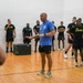 A civilian instructor or trainer in a blue shirt is addressing a group of U.S. Army soldiers wearing black 'ARMY' branded physical training uniforms inside what appears to be a gymnasium or racquetball court. The instructor appears to be demonstrating or explaining something with hand gestures while the soldiers stand attentively around him. A black plyo box is visible on the hardwood floor, suggesting a physical fitness or training session.