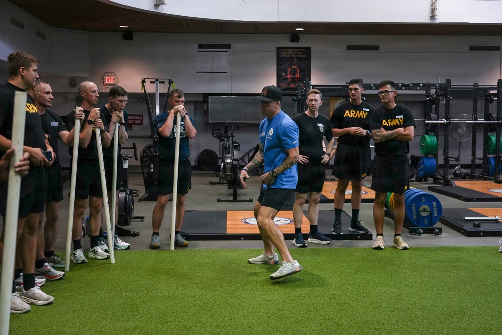 A fitness instructor or coach wearing a blue shirt and black cap is demonstrating a movement or exercise technique on an artificial turf surface inside a modern military gym facility. A group of individuals, several wearing black 'ARMY' branded athletic uniforms, observe attentively while some lean on long poles or bars. The setting appears to be a U.S. Army strength and conditioning training session.
