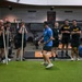A fitness instructor or coach wearing a blue shirt and black cap is demonstrating a movement or exercise technique on an artificial turf surface inside a modern military gym facility. A group of individuals, several wearing black 'ARMY' branded athletic uniforms, observe attentively while some lean on long poles or bars. The setting appears to be a U.S. Army strength and conditioning training session.