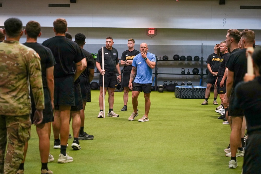 A group of military personnel in Army PT uniforms and one individual in camouflage are gathered in a modern indoor fitness facility. A civilian instructor or trainer in a light blue shirt appears to be briefing or instructing the group, who are arranged in a semicircle on artificial turf. The setting suggests a physical training session or fitness demonstration.