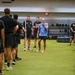 A group of military personnel in Army PT uniforms and one individual in camouflage are gathered in a modern indoor fitness facility. A civilian instructor or trainer in a light blue shirt appears to be briefing or instructing the group, who are arranged in a semicircle on artificial turf. The setting suggests a physical training session or fitness demonstration.