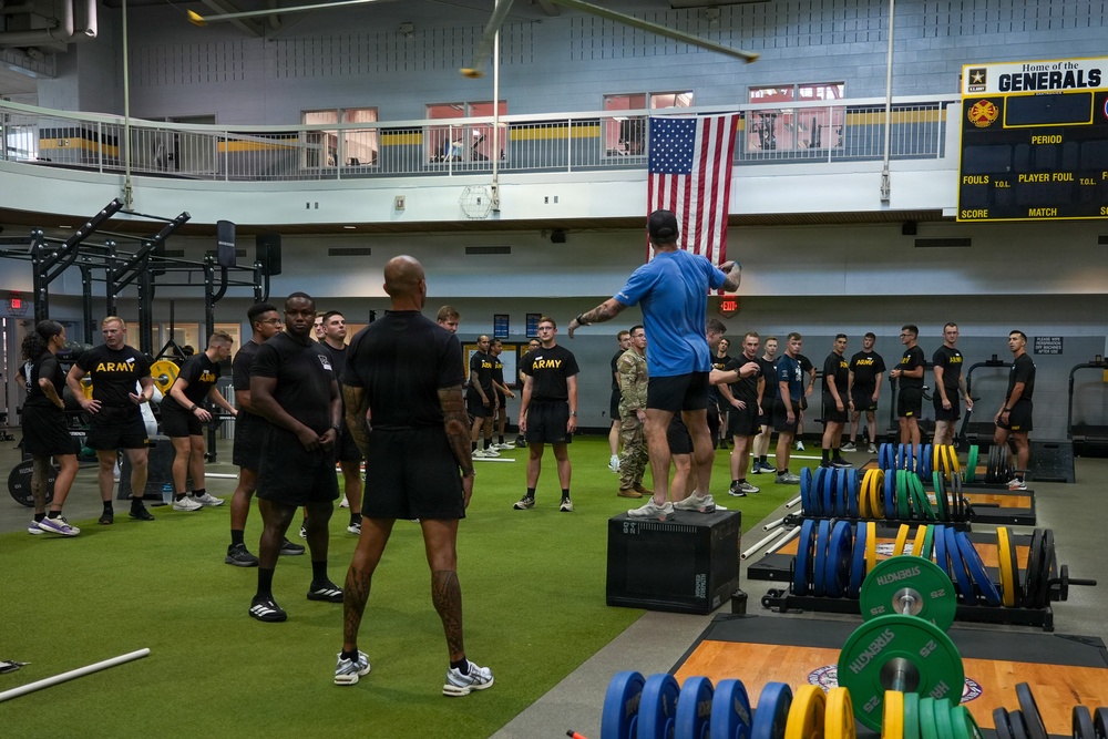 A large group of U.S. Army personnel in black PT (physical training) uniforms are gathered in a military fitness facility. An instructor in a blue shirt is standing elevated on a plyo box, appearing to brief or instruct the group. The facility features weightlifting equipment, a scoreboard reading 'Home of the Generals,' and a large American flag hanging on the wall.