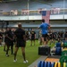 A large group of U.S. Army personnel in black PT (physical training) uniforms are gathered in a military fitness facility. An instructor in a blue shirt is standing elevated on a plyo box, appearing to brief or instruct the group. The facility features weightlifting equipment, a scoreboard reading 'Home of the Generals,' and a large American flag hanging on the wall.