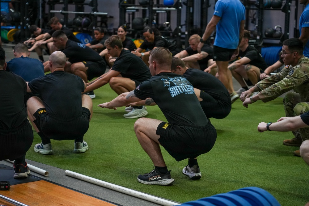 A large group of military personnel are performing squat exercises on an artificial turf surface inside a modern fitness facility. The majority are wearing black Army PT (Physical Training) uniforms, while at least one individual is in Army Combat Uniform (ACU) and another in a blue shirt. The session appears to be a structured group physical training event.