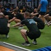A large group of military personnel are performing squat exercises on an artificial turf surface inside a modern fitness facility. The majority are wearing black Army PT (Physical Training) uniforms, while at least one individual is in Army Combat Uniform (ACU) and another in a blue shirt. The session appears to be a structured group physical training event.