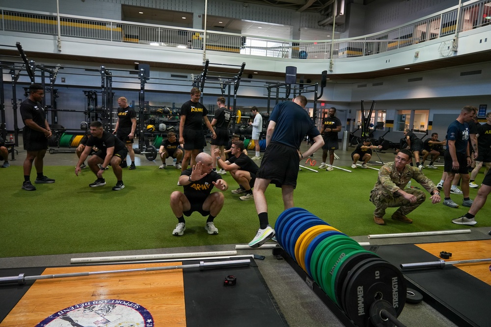 A large group of U.S. Army soldiers and personnel are conducting a physical training session inside a modern military fitness facility. The participants are performing what appears to be agility or warm-up drills on an artificial turf area, supervised by instructors. The facility is well-equipped with barbells, weight racks, and resistance training equipment.