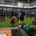 A large group of U.S. Army soldiers and personnel are conducting a physical training session inside a modern military fitness facility. The participants are performing what appears to be agility or warm-up drills on an artificial turf area, supervised by instructors. The facility is well-equipped with barbells, weight racks, and resistance training equipment.