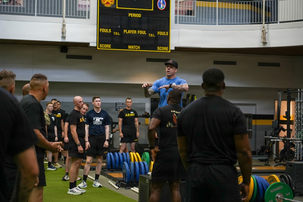 A fitness instructor or trainer in a blue shirt stands elevated, demonstrating or explaining an exercise to a large group of military personnel in a gymnasium. The group, wearing predominantly black Army-branded athletic wear, is gathered in a semicircle listening to the instruction. The setting appears to be a military fitness facility equipped with barbells, weight plates, and training equipment.