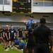 A fitness instructor or trainer in a blue shirt stands elevated, demonstrating or explaining an exercise to a large group of military personnel in a gymnasium. The group, wearing predominantly black Army-branded athletic wear, is gathered in a semicircle listening to the instruction. The setting appears to be a military fitness facility equipped with barbells, weight plates, and training equipment.