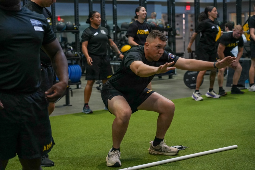 A group of individuals wearing U.S. Army physical training uniforms (black shirts with 'ARMY' text and gold lettering) are exercising in a modern military fitness facility. The central figure is performing a squat or lateral movement exercise near a white PVC pipe on an artificial turf floor. The facility is well-equipped with barbells, weight racks, and bumper plates visible in the background.