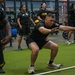 A group of individuals wearing U.S. Army physical training uniforms (black shirts with 'ARMY' text and gold lettering) are exercising in a modern military fitness facility. The central figure is performing a squat or lateral movement exercise near a white PVC pipe on an artificial turf floor. The facility is well-equipped with barbells, weight racks, and bumper plates visible in the background.
