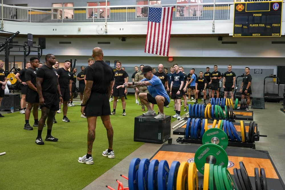 A large group of U.S. Army personnel in black 'ARMY' branded athletic wear are gathered in a military fitness facility or gymnasium. An instructor in a blue shirt is demonstrating a box squat or box jump exercise on a plyo box while others observe. The facility is well-equipped with weight plates, barbells, and exercise equipment, and features a large American flag on the wall.