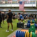 A large group of U.S. Army personnel in black 'ARMY' branded athletic wear are gathered in a military fitness facility or gymnasium. An instructor in a blue shirt is demonstrating a box squat or box jump exercise on a plyo box while others observe. The facility is well-equipped with weight plates, barbells, and exercise equipment, and features a large American flag on the wall.
