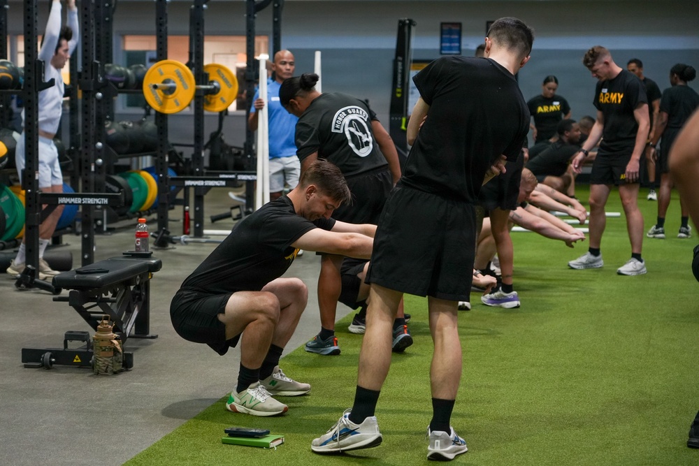 A group of U.S. Army soldiers and National Guard personnel are conducting a physical training session inside a military gymnasium. The participants, dressed in black Army PT uniforms, are performing stretching and warm-up exercises in an organized formation. The facility is well-equipped with weightlifting racks, Hammer Strength equipment, and turf flooring.