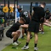 A group of U.S. Army soldiers and National Guard personnel are conducting a physical training session inside a military gymnasium. The participants, dressed in black Army PT uniforms, are performing stretching and warm-up exercises in an organized formation. The facility is well-equipped with weightlifting racks, Hammer Strength equipment, and turf flooring.